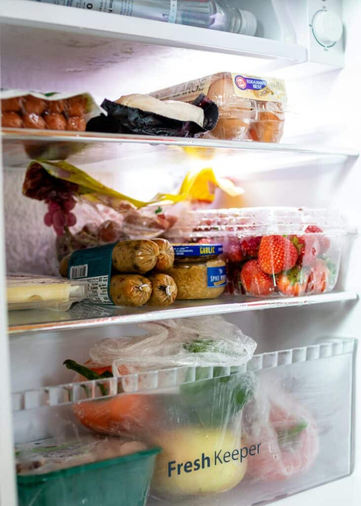 Brightly lit open refrigerator with eggs, meats, fruits and vegetables inside.